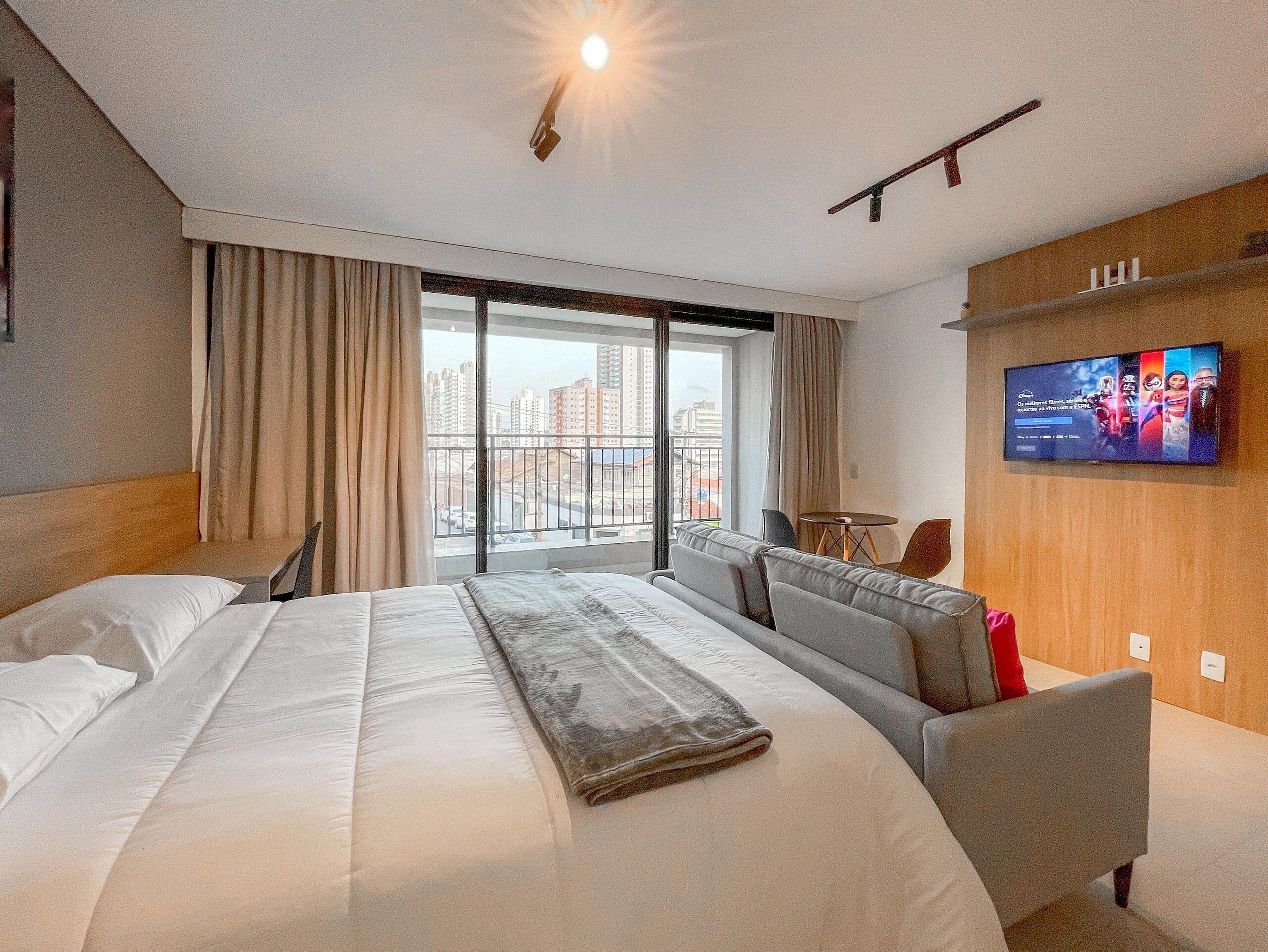 A hotel room with a large bed, a sofa, a TV, and a view of the city.