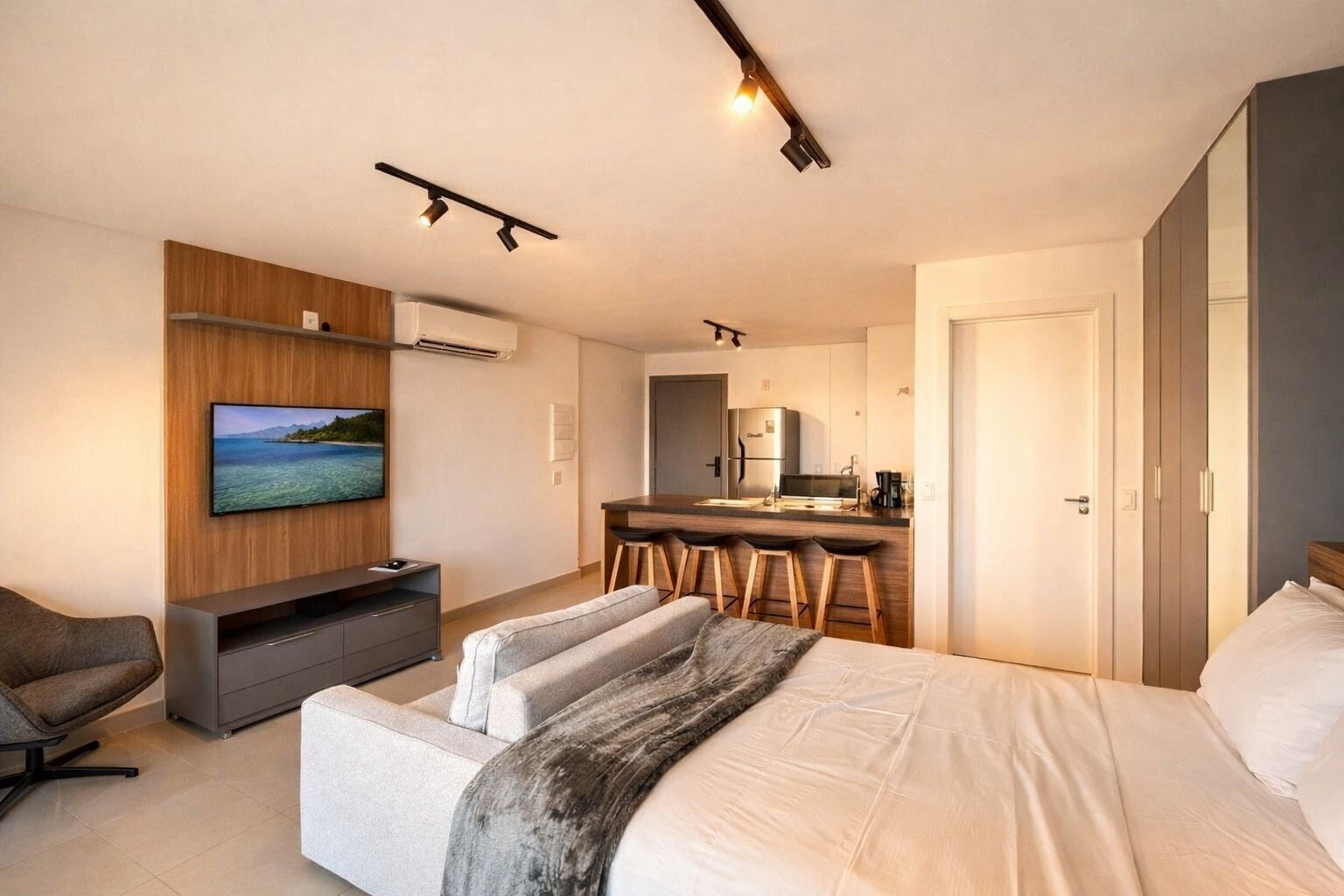 A modern hotel room with a bed, a TV mounted on a wooden panel, a dining area with stools, and a kitchenette.
