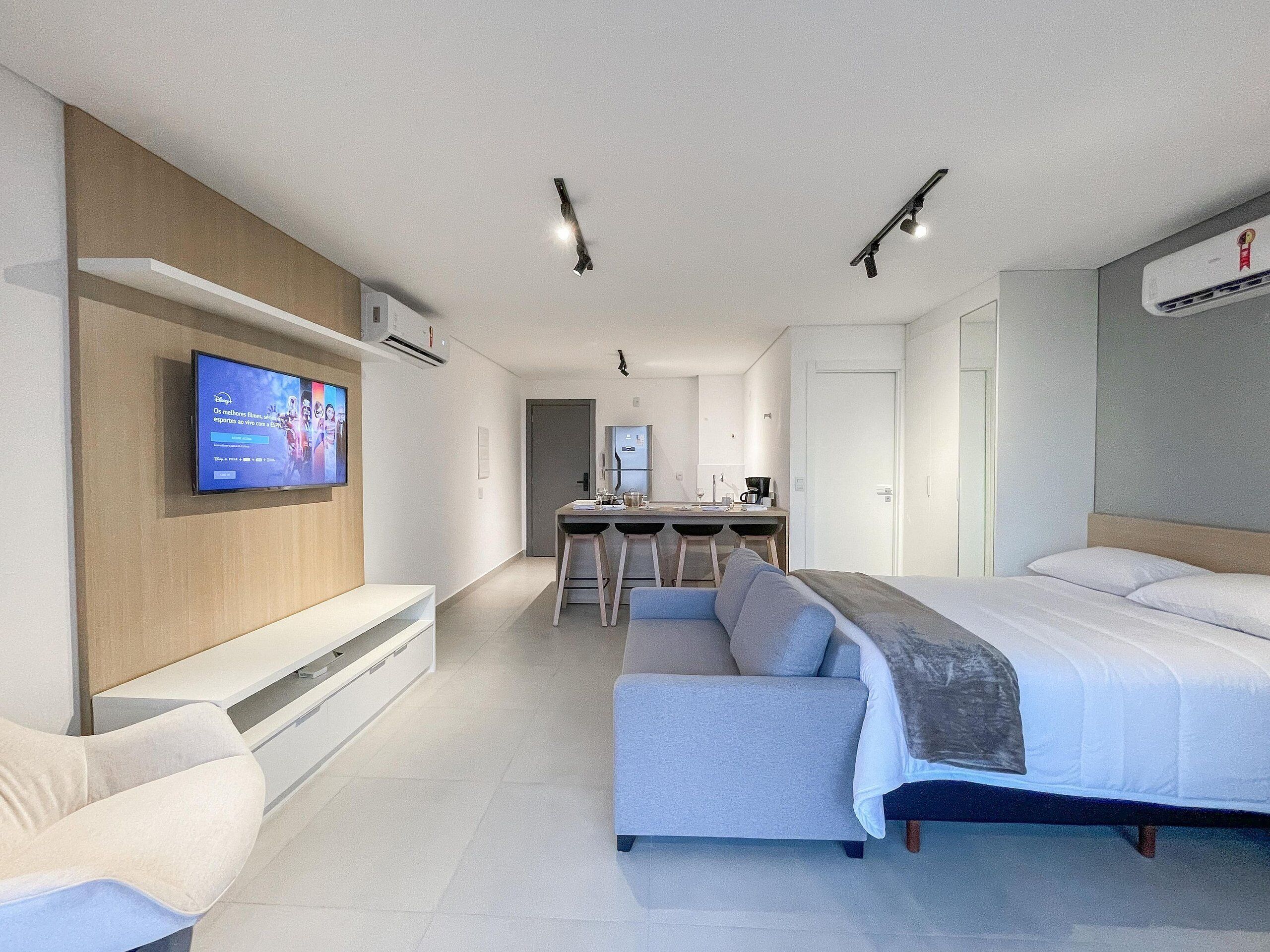A modern hotel room with a sofa, a bed, a TV mounted on the wall, a kitchenette, and a ceiling light.