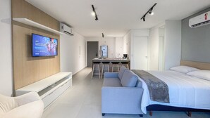 A modern hotel room with a sofa, a bed, a TV mounted on the wall, a kitchenette, and a ceiling light.