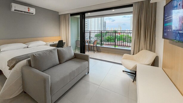 A modern hotel room with a bed, a sofa, a desk, and a balcony with a city view.