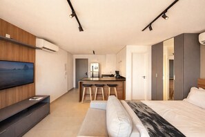 A modern hotel room with a bed, a TV, a kitchen area, and a seating area.