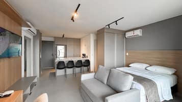 A modern hotel room with a sofa, a bed, a desk, and a kitchenette.