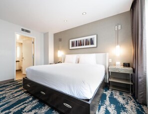 Ein Hotelzimmer mit einem großen Bett, einem Schreibtisch mit Lampe, einem Bild an der Wand und einem durchgehenden Türspalt einsehbarer Badezimmer.