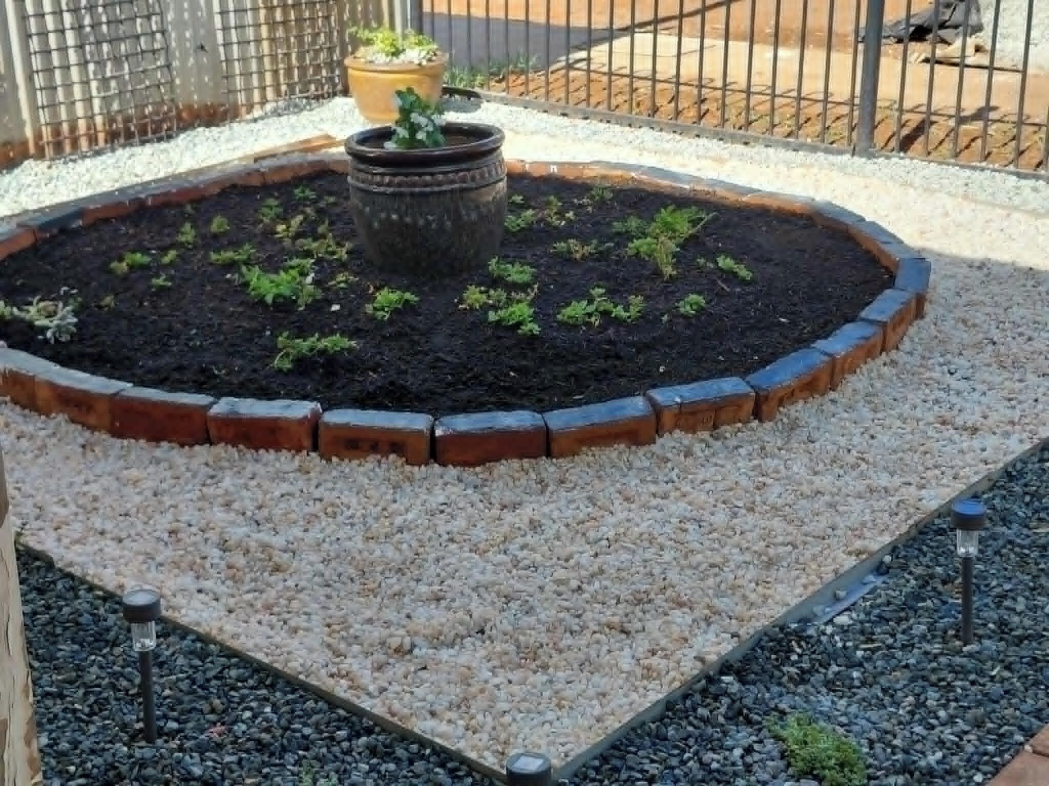 Cama de jardín circular con borde de ladrillo y piedras, rodeada por una valla metálica.