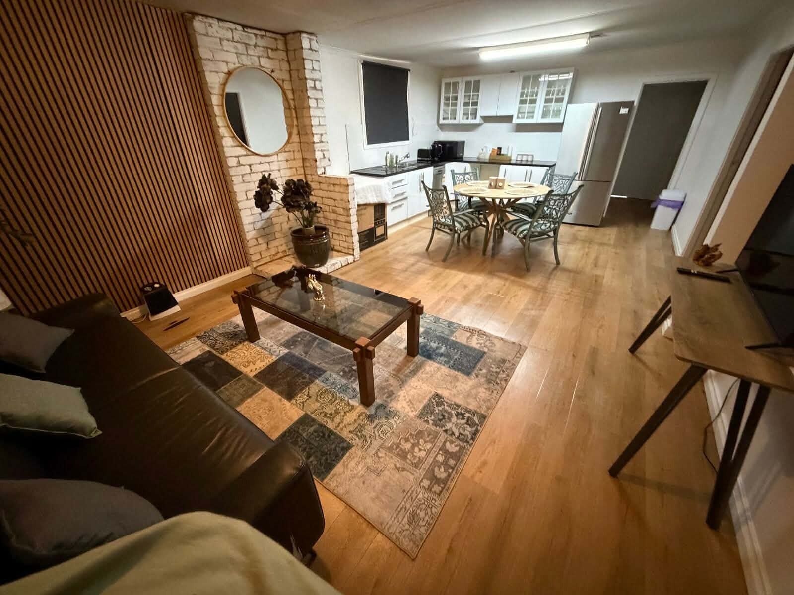 A living room with a sofa, coffee table, and a rug. There is a kitchen area with cabinets and a dining table with chairs.