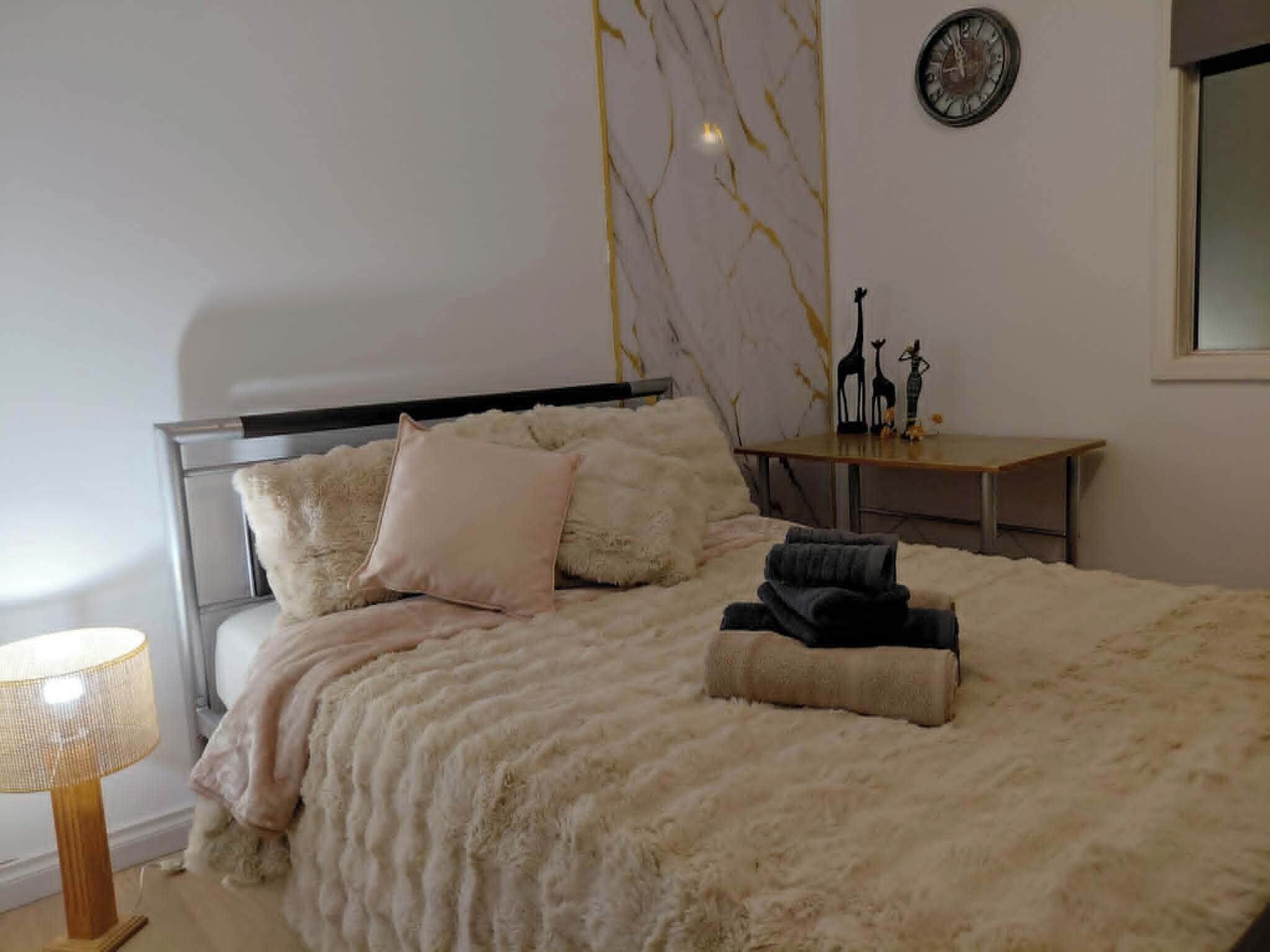 A bedroom with a bed, pillows, a bedside table with decorative items, a lamp, and a wall clock.