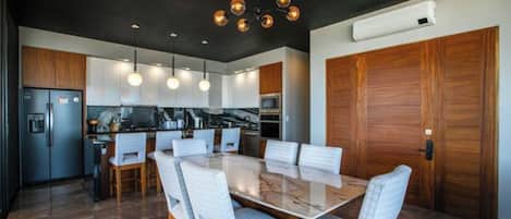 A modern kitchen with a dining area, featuring a marble table, white upholstered chairs, and pendant lighting.