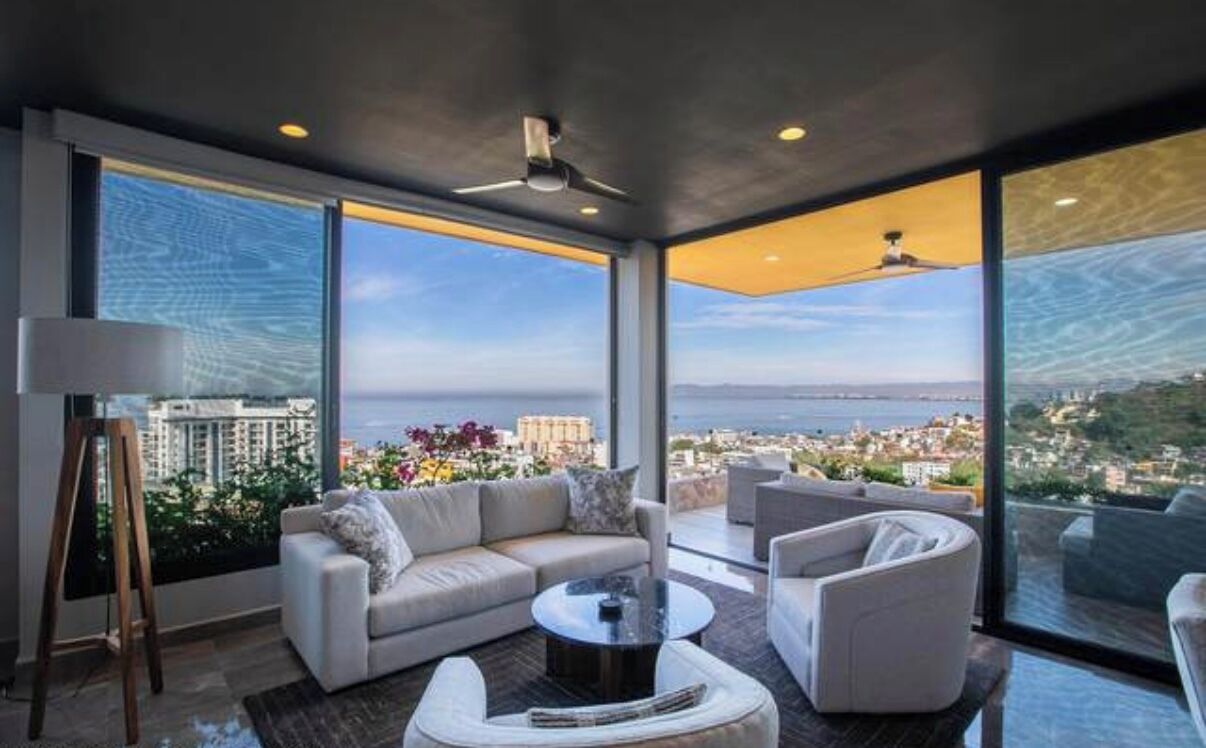 Un salon moderne comprenant un canapé, des fauteuils et une table basse, avec de grandes fenêtres offrant une vue sur la ville.