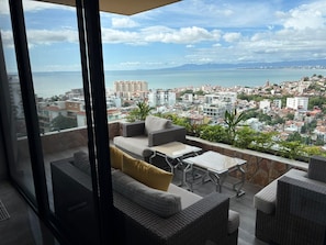 A balcony with wicker furniture, a view of a coastal cityscape, and a clear sky. - Casa Mirador Luxury 3 bedroom Condo, Panoramic Ocean Views (Puerto Vallarta)