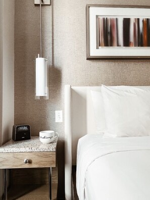 Een hotelkamer met een bed, een nachtkastje met een koffiekop en een klok, een hanglamp en een lijst met een abstract schilderij aan de muur.