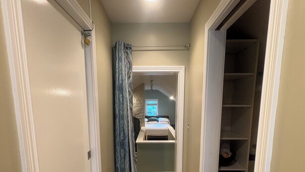 Un couloir étroit, dont le sol est recouvert de moquette, mène à une chambre à coucher équipée d’un lit et d’une fenêtre.