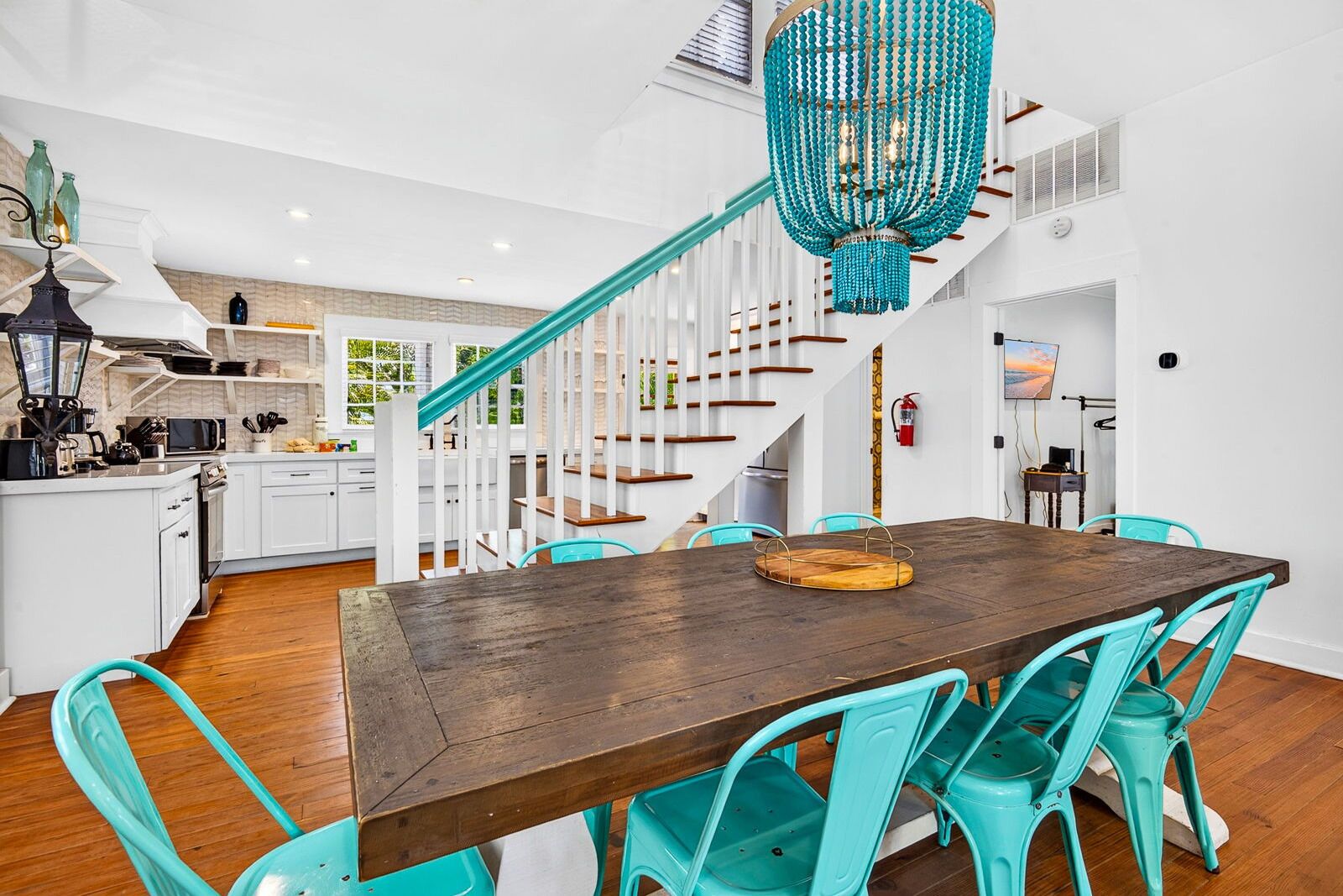 A modern kitchen with white cabinetry, a wooden dining table, turquoise chairs, and a staircase with white railings.