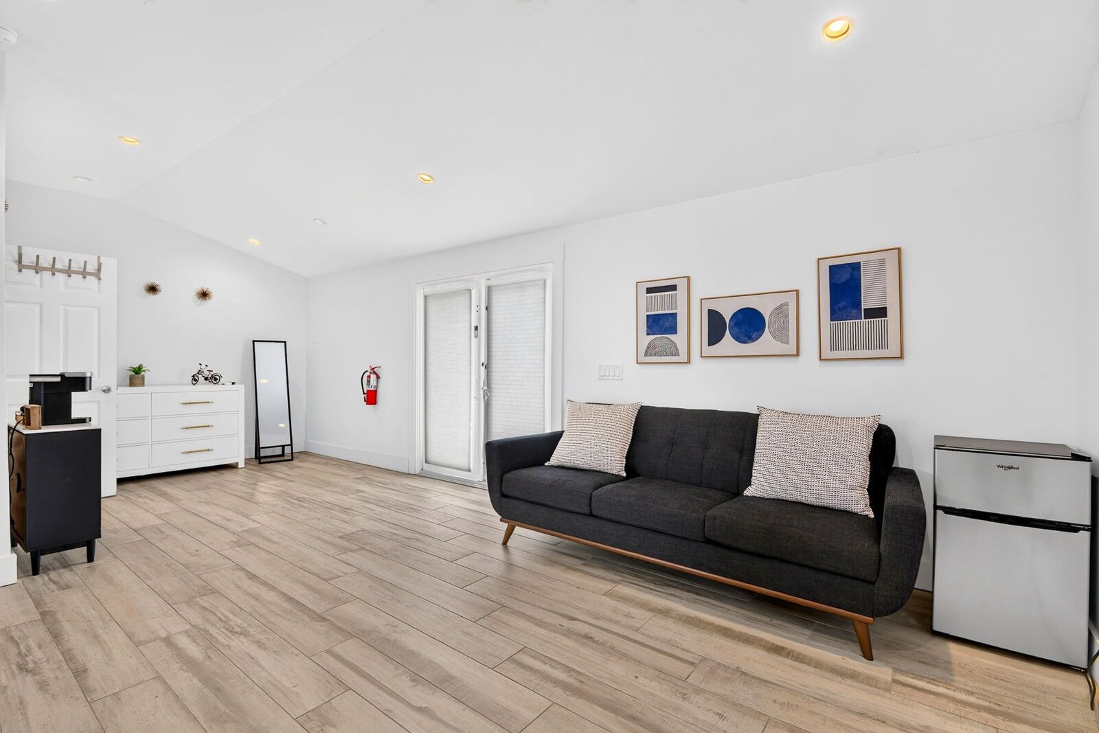 Un salon moderne avec un canapé gris, une petite kitchenette et une porte blanche.