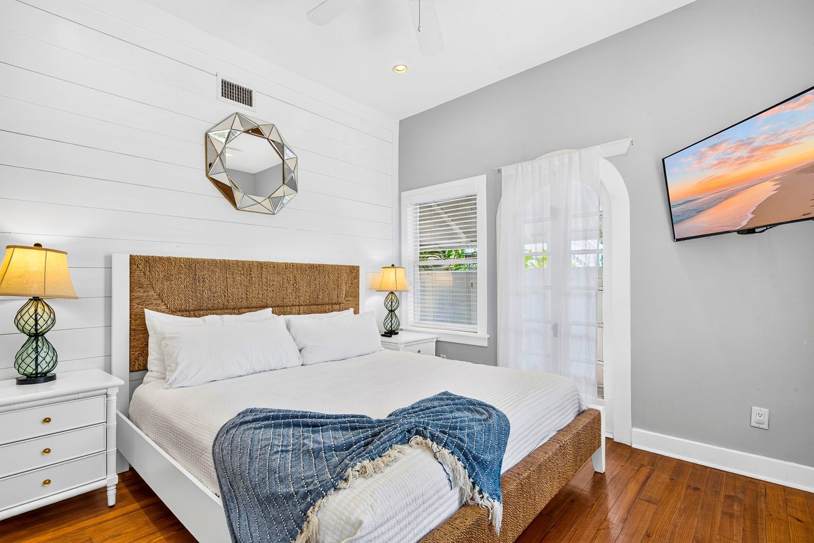 Une chambre à coucher comprenant un lit, deux tables de chevet, un téléviseur fixé au mur, une fenêtre avec des rideaux et un miroir décoratif.