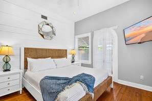 Ein Schlafzimmer mit einem Bett, zwei Nachttischen, einem an der Wand befestigten Fernseher, einem Fenster mit Vorhängen und einem dekorativen Spiegel.