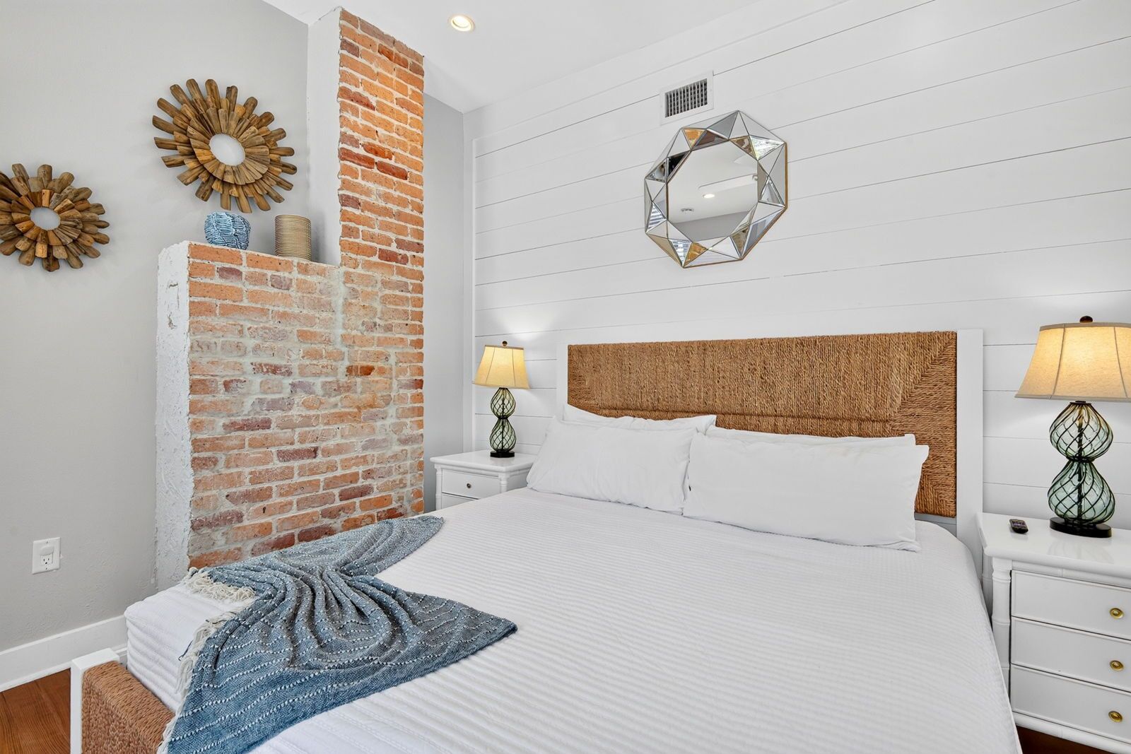 A bedroom with a brick accent wall, a bed with white linens, two bedside tables with lamps, and a decorative mirror on the wall.