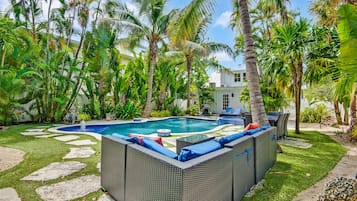 A swimming pool with a surrounding garden, a lounge area with wicker furniture, and a white building in the background.