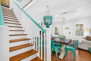 Un escalier avec des marches en bois et des rampes blanches, un coin repas avec une table en bois et des chaises de couleur turquoise, et un lustre avec un abat-jour turquoise.