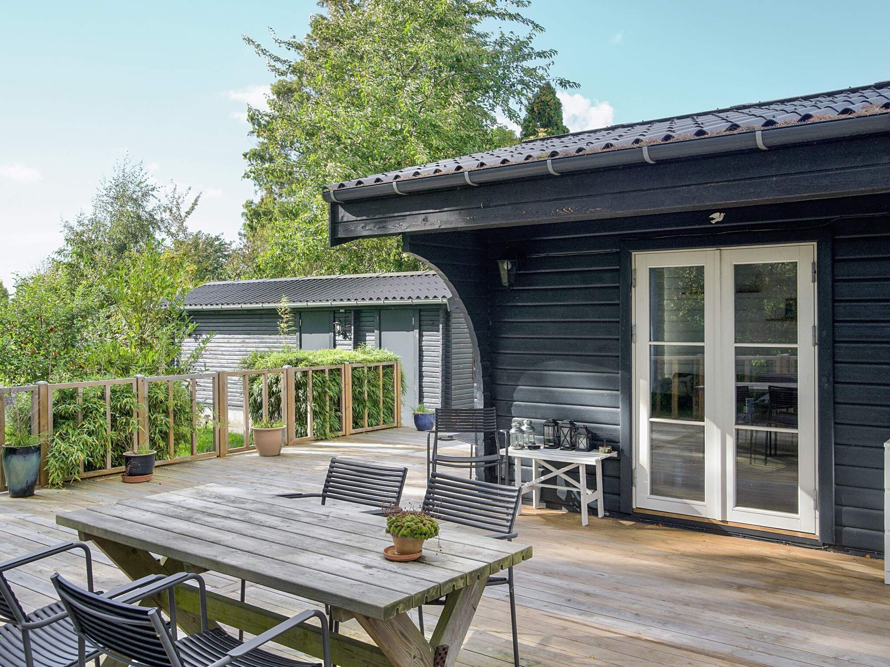 A wooden deck with a table and chairs, a potted plant, and a building with a dark exterior and large glass doors.