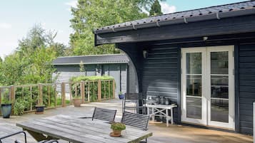 Eine Holzterrasse mit Tisch und Stühlen, einem Blumentopf und einem Gebäude mit dunkler Fassade und großen Glasüren.