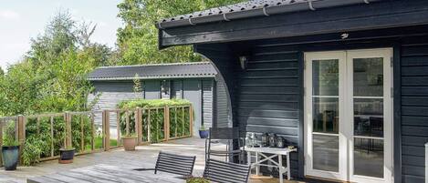 A wooden deck with a table and chairs, a potted plant, and a building with a dark exterior and large glass doors.