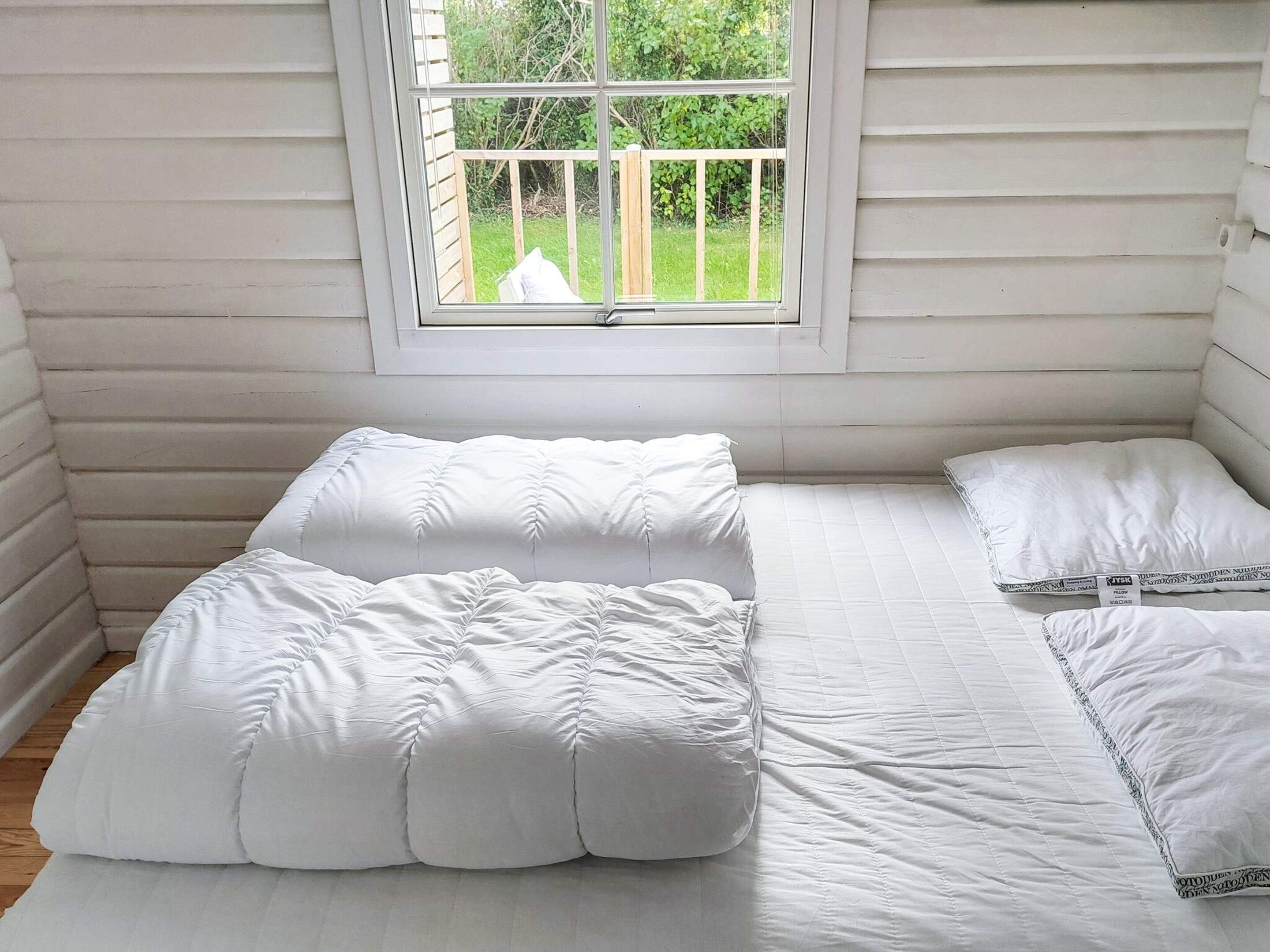 Ein kleiner Raum mit weißen Paneelen, einem Bett, einem Fenster und Blick auf eine Holzterrasse.