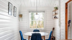 Uma  área de refeições com uma mesa redonda, cadeiras azuis, um violão e uma janela com vista para o verde.