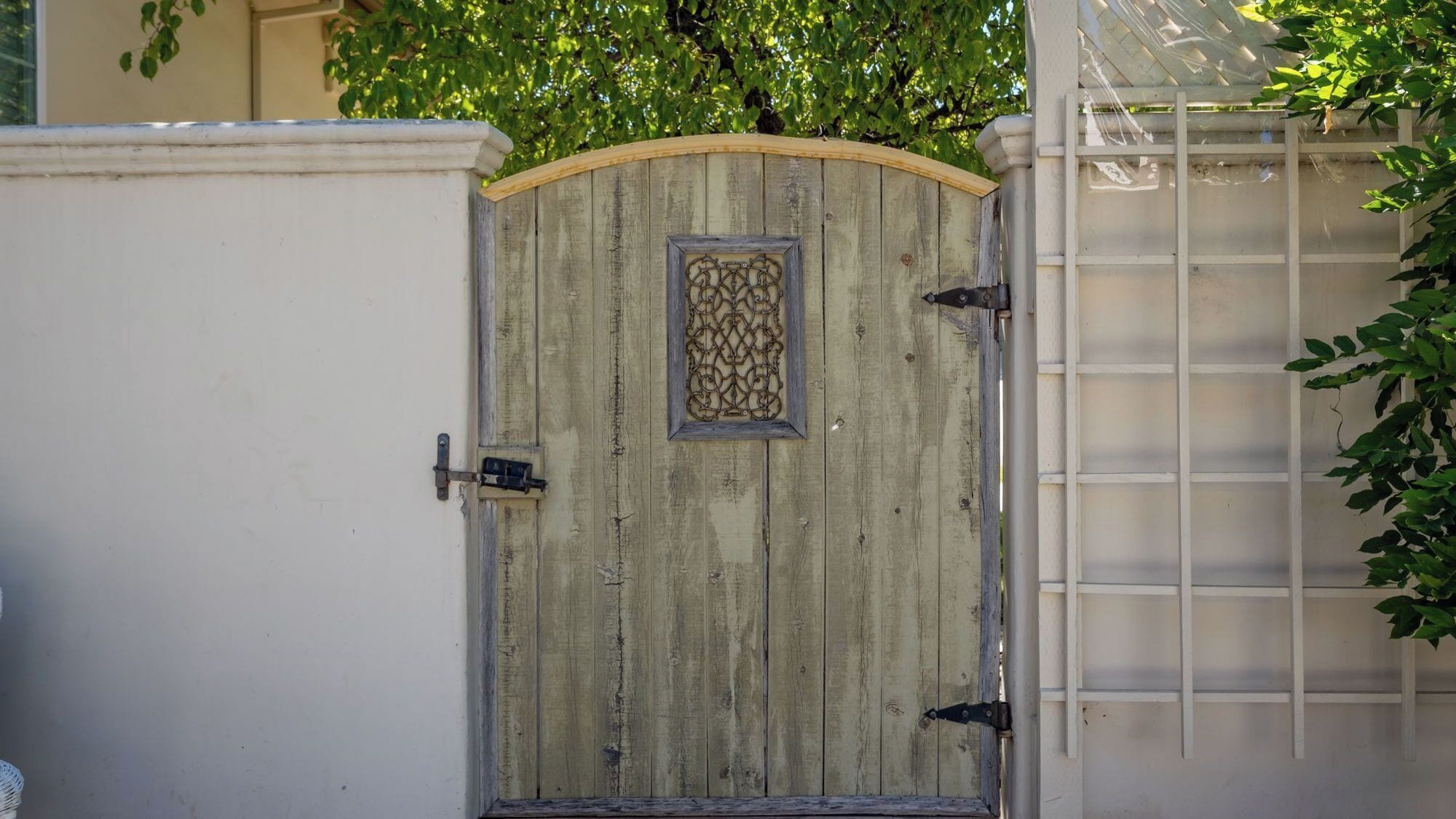 A wooden gate with a decorative metal frame, a latch, and a white wall with a window to the left.