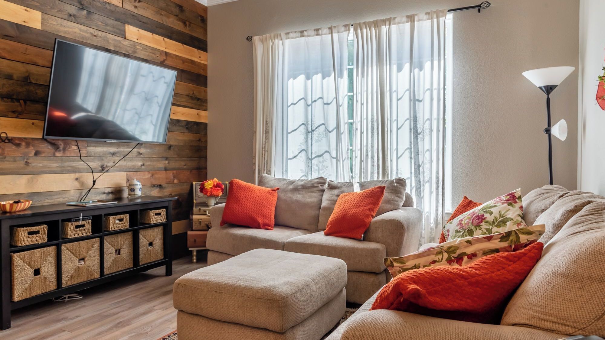 Un salon avec un téléviseur à écran plat fixé à un mur en bois, un ensemble de canapés avec des coussins orange, une table d’appoint et un lampadaire.