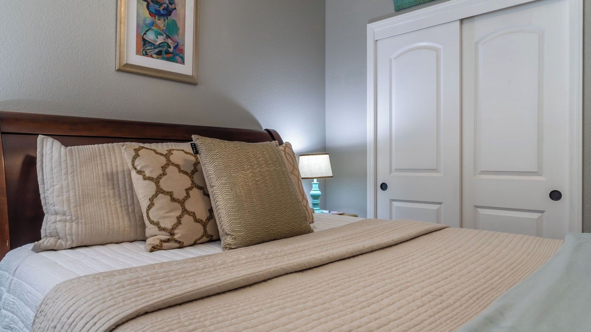 Een net opgemaakt bed met een beige deken en twee decoratieve kussens, een witte deur en een aan de muur hangend schilderij.