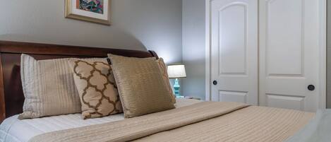 Een net opgemaakt bed met een beige deken en twee decoratieve kussens, een witte deur en een aan de muur hangend schilderij.