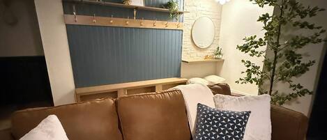A brown leather sofa with white and patterned cushions, a wooden shelf with decorative items, a round mirror, and a potted plant.