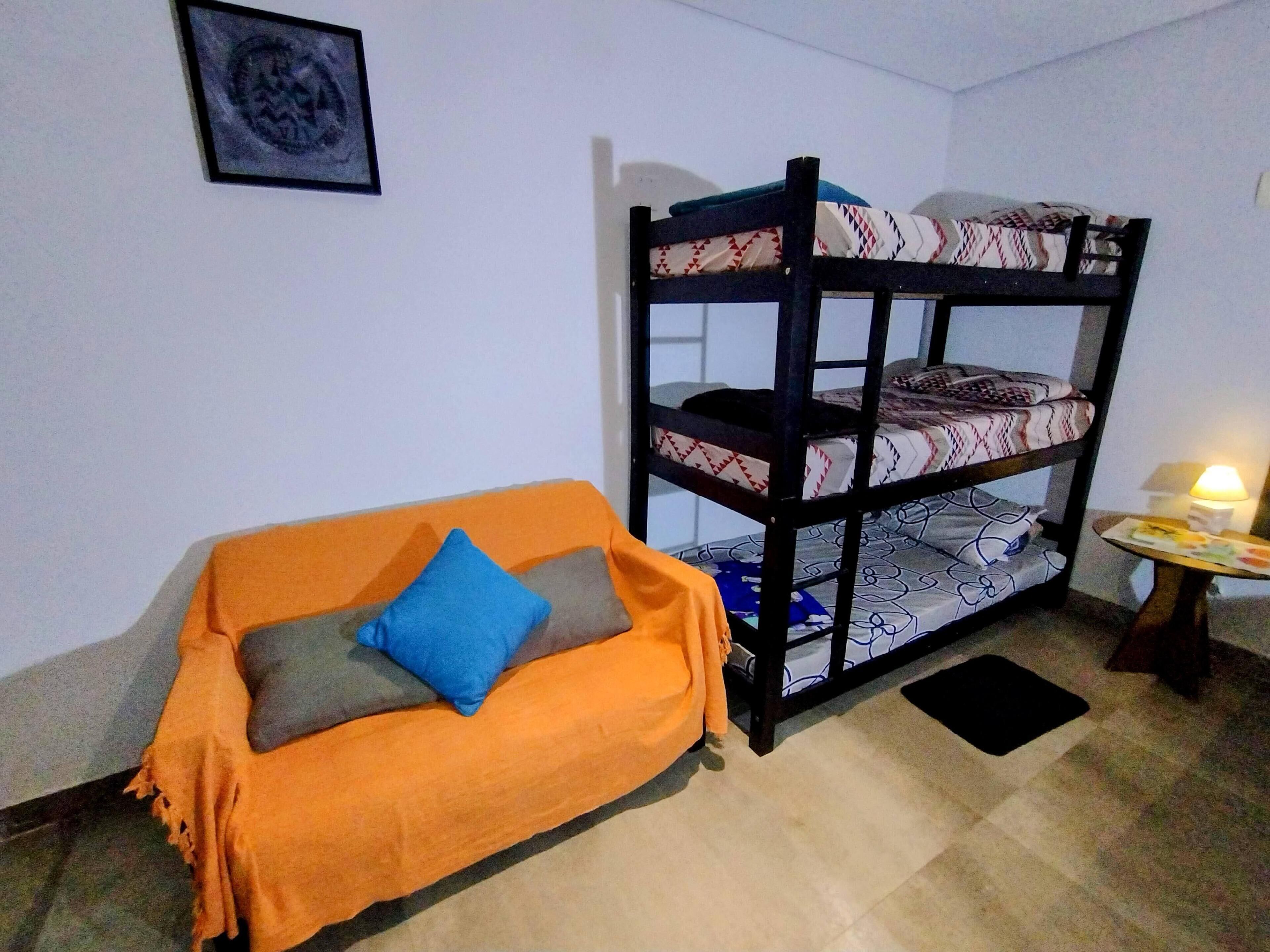 Ein Stockbett mit gemusterten Betttextilien, ein orangefarbenes Sofa mit Kissen, ein kleiner Tisch mit einer Lampe und ein gerahmtes Bild an der Wand.