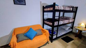 Ein Stockbett mit gemusterten Betttextilien, ein orangefarbenes Sofa mit Kissen, ein kleiner Tisch mit einer Lampe und ein gerahmtes Bild an der Wand.