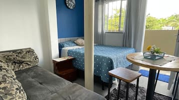 Een kamer met een grijze bank, een bed met blauw beddengoed, een klein tafeltje met een mand en een raam met gordijnen.