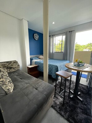 A room with a grey sofa, a bed with blue bedding, a small table with a basket, and a window with curtains.