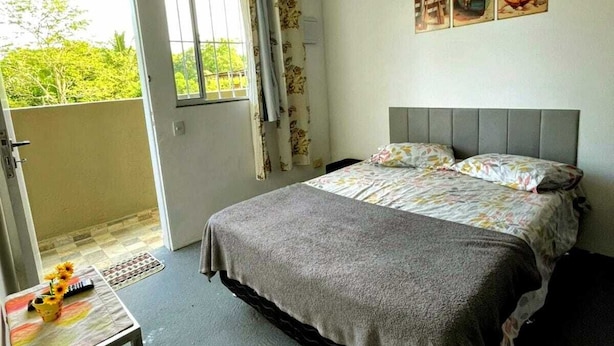 A bedroom with a bed, a small table with a vase, and a view of greenery outside.