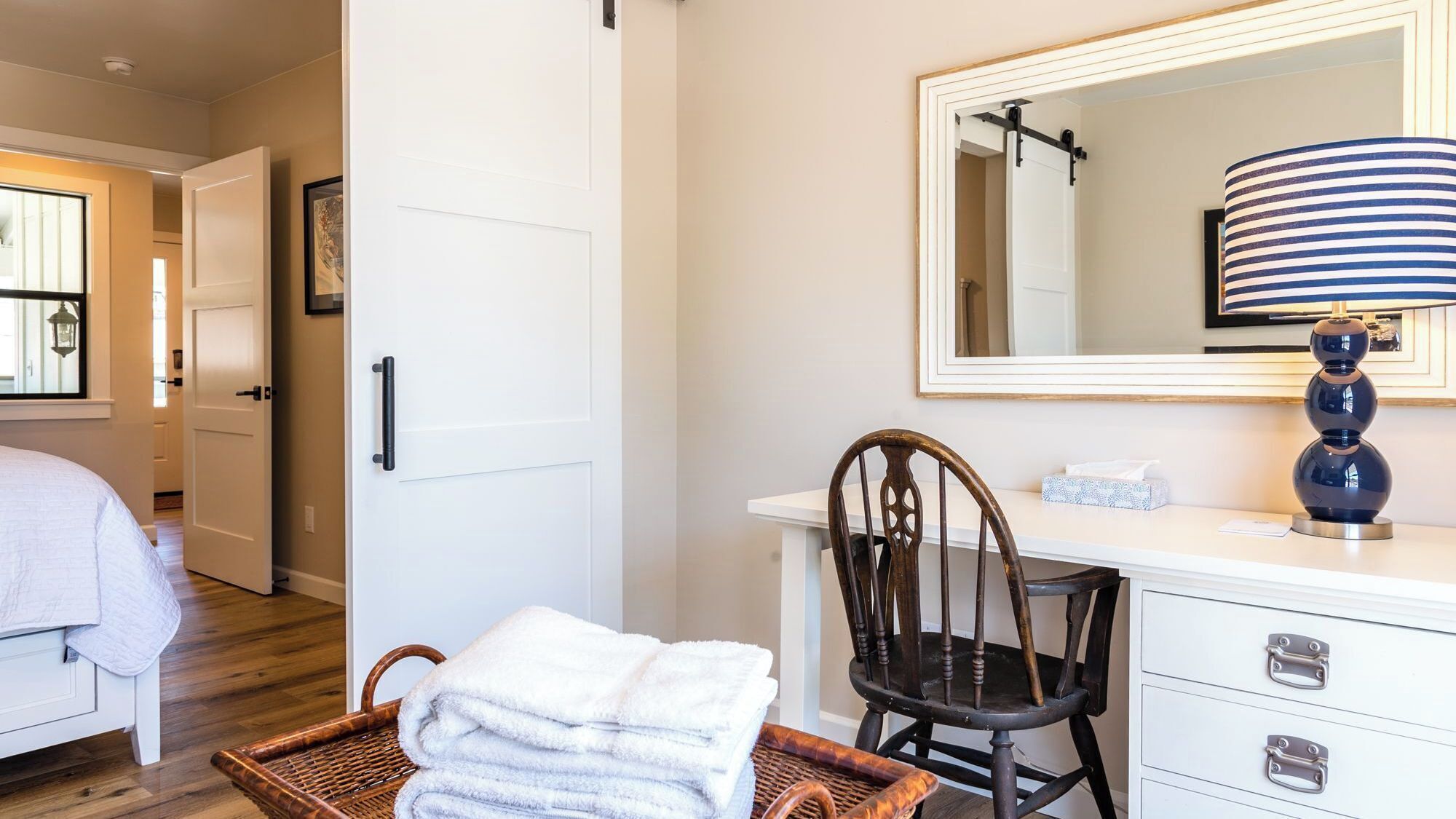 A hotel room with a bed, a chair, a desk with a lamp, a mirror, and a basket of towels.