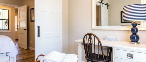 A hotel room with a bed, a chair, a desk with a lamp, a mirror, and a basket of towels.