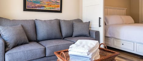 A grey sofa with multiple cushions, a wicker basket with folded towels, and a bed with white bedding.