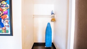 A narrow hallway with a framed Mario poster on the left, a blue ironing board in the center, and a white shelf with a blue object and yellow items on the right. - Accessible King Room (Anaheim)