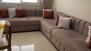 A living room with a sectional sofa, a dining table, and a window with blinds.