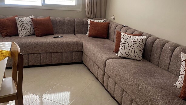 A living room with a sectional sofa, a dining table, and a window with blinds. - Appartement lumineux calme et bien situé á 2 pas de la plage-Skhirat et Sablette (Maroc)
