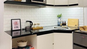 Een compacte keuken met witte kastjes, een zwart aanrecht, een magnetron en een koelkast. Er staan potplanten op de planken en een lijst op het aanrecht.