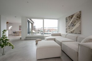 A modern living room with a large window, a sofa, a dining area, and a painting on the wall.
