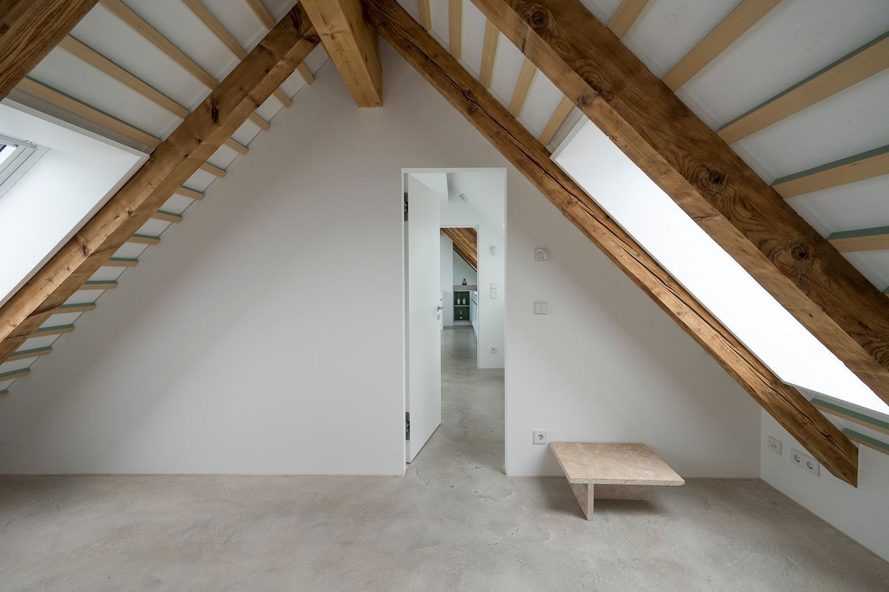 Ein Dachzimmer mit hohen Decken, freiliegenden Holzbalken, einem Betonboden und einer kleinen Bank.