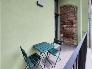 Un balcón con una mesa verde y dos sillas, una rústica puerta de madera y una pequeña lámpara en la pared.
