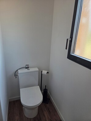 A small, compact bathroom with a white toilet, a black toilet paper holder, and a window with a frosted glass panel. - 130-square-meter Villa in Montalivet, 4 bedrooms, near the ocean (Vensac)