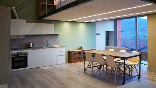 A modern kitchen with a dining area, wooden flooring, and a view of the cityscape through large windows. - Ronchetto - Appartamento Atticus (Cunardo)
